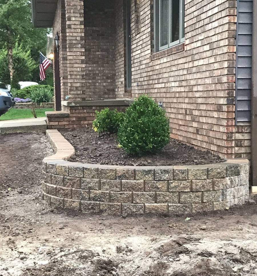 A curved block retaining wall at the corner of a brick house, with a boxwood behind it and bare soil in front.