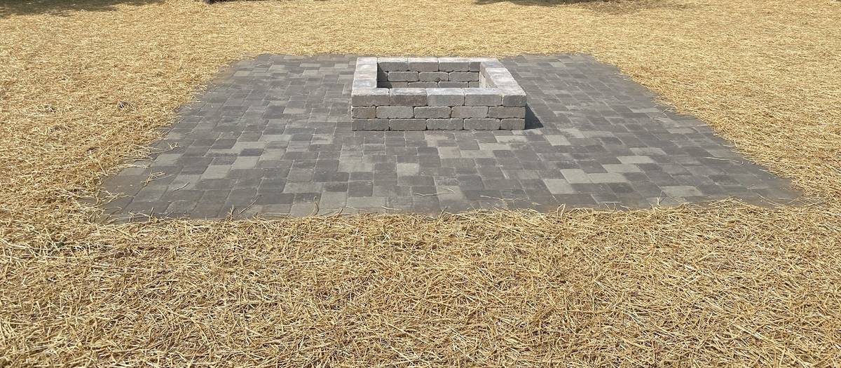 A gray paver patio with a square block fire pit in the middle of a fenced backyard, straw covering the ground around it.
